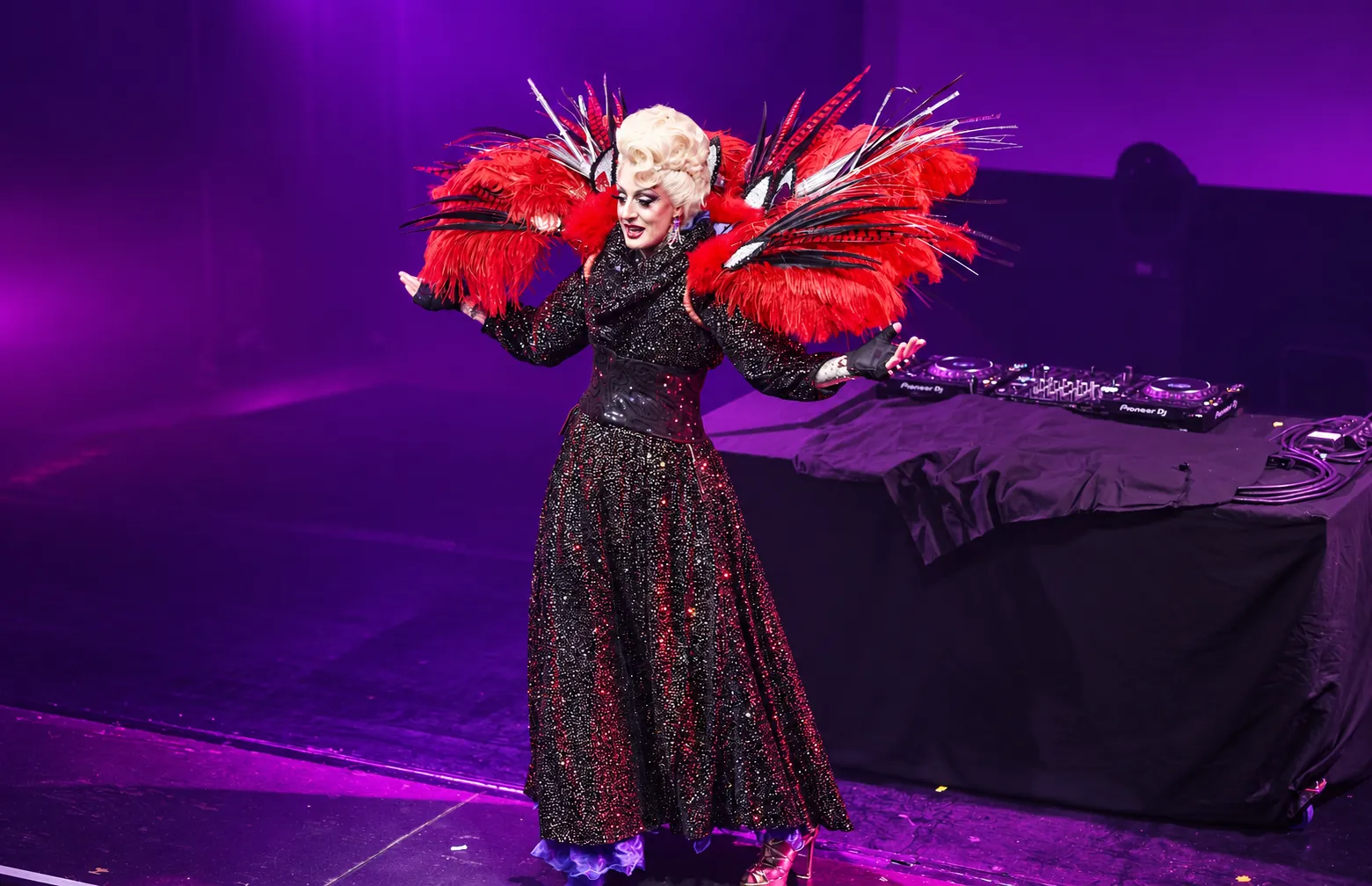 Lady Maxx Paris sur scène en costume à sequins et grandes plumes rouges, show drag queen sous lumières violettes