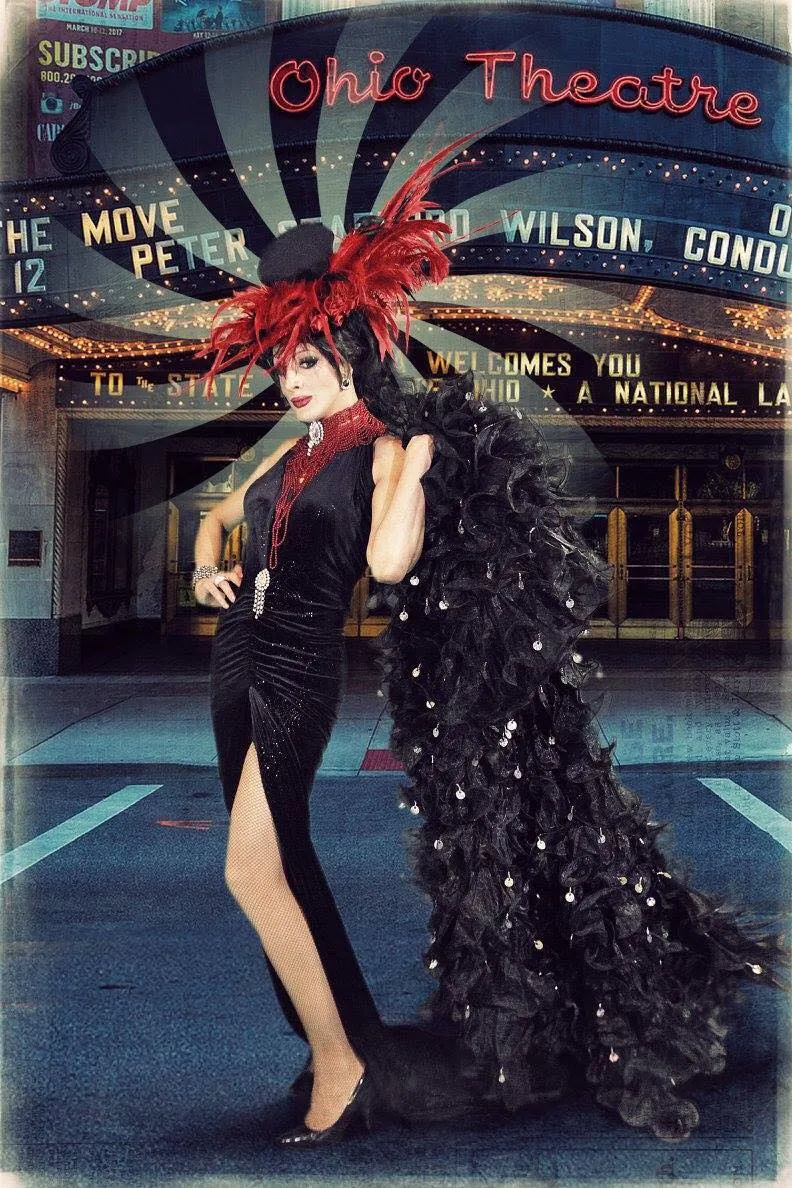 Lady Maxx, drag queen cabaret en robe noire fendue, chapeau à plumes rouges et boa à sequins devant un théâtre