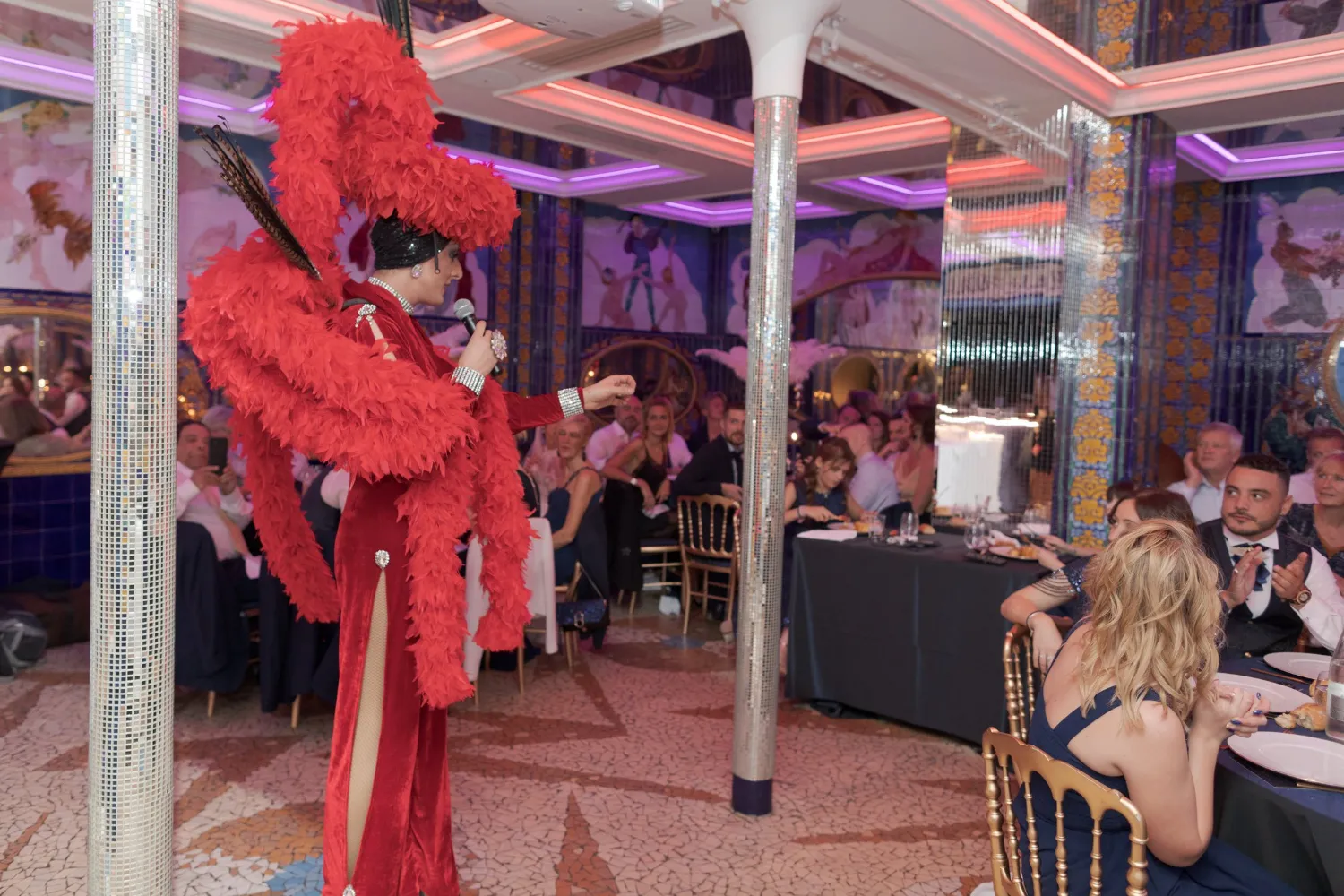 Lady Maxx en plumes rouges au micro face aux invités d'un dîner-spectacle à Paris