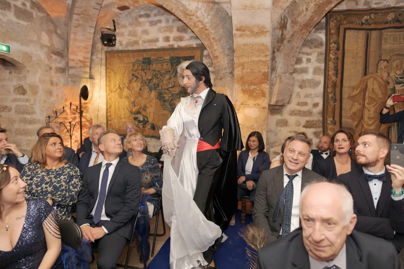 Lady Maxx transformiste au milieu des invités d'un mariage dans une salle voûtée